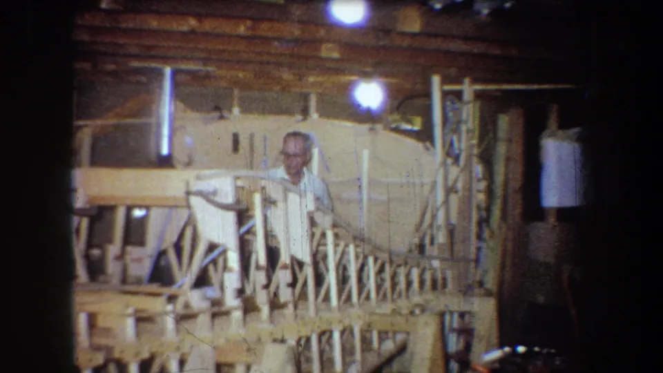 The older man looks toward the camera from behind the large wooden structure, with workshop supports and overhead lighting visible around him.