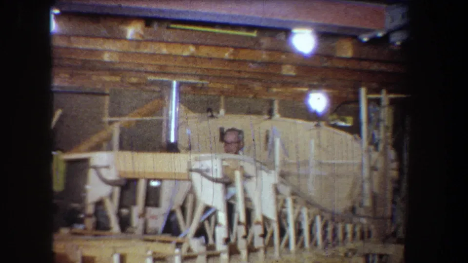 1965 home-workshop frame showing a man in a light shirt behind handmade wooden framing under warm indoor lighting.