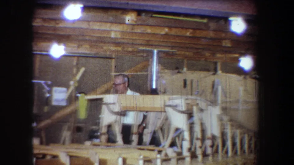 1965 Yucaipa home-movie frame of a man behind wooden framing in a private workshop with exposed bulbs and paneled walls.
