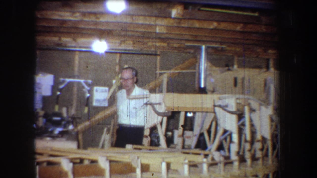 1965 Yucaipa home-movie frame showing a man in a white shirt near a wooden boat structure in a cluttered interior workshop.