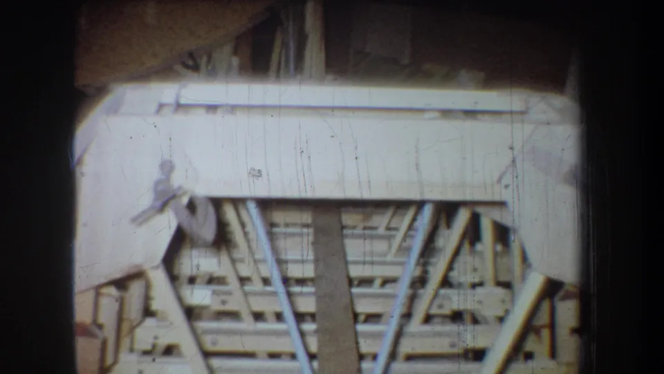 Close 1965 home-movie view of an unfinished wooden boat section, showing pale planks, ribs and support framing in the workshop.