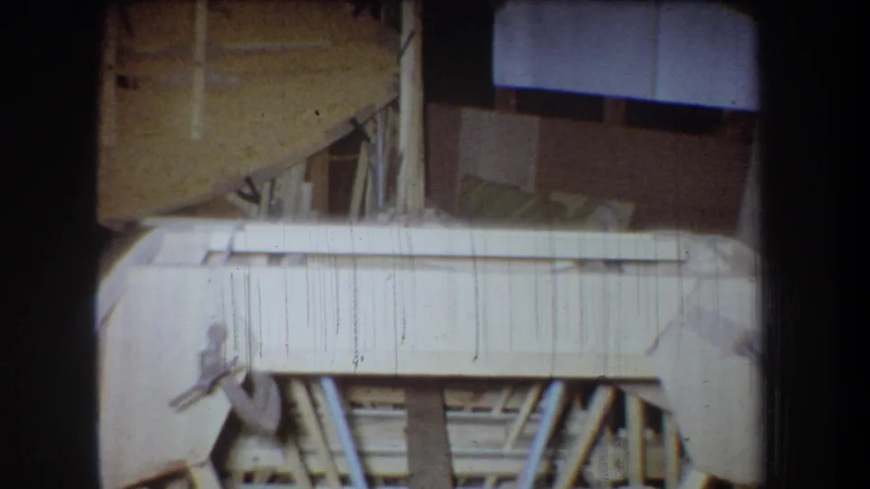 1965 Yucaipa home-movie frame showing a close view of the wooden boat structure, braces and interior workshop background.