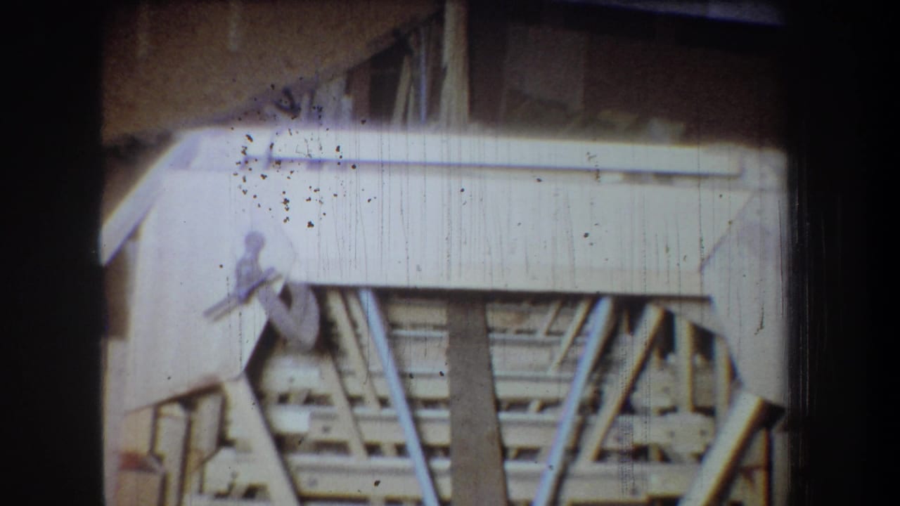 1965 8mm frame from Volmer Yucaipa showing unfinished attic rafters, pale boards, and dark surrounding film edges.