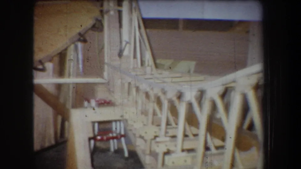 1965 Yucaipa home-movie frame showing a side section of unfinished wooden airplane framing inside a workshop-like space.