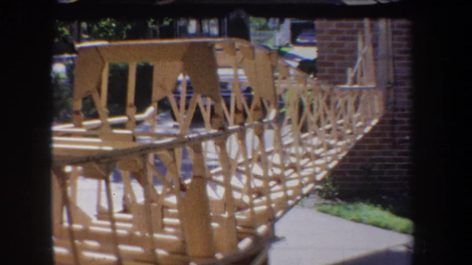 Wide 1965 Yucaipa home-movie frame of a large handmade wooden model with truss railings, set beside brickwork and greenery.