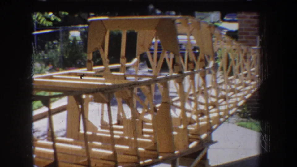 1965 8mm frame from Yucaipa showing the handmade wooden model from the side, with repeated braces, deck boards, and outdoor surroundings.