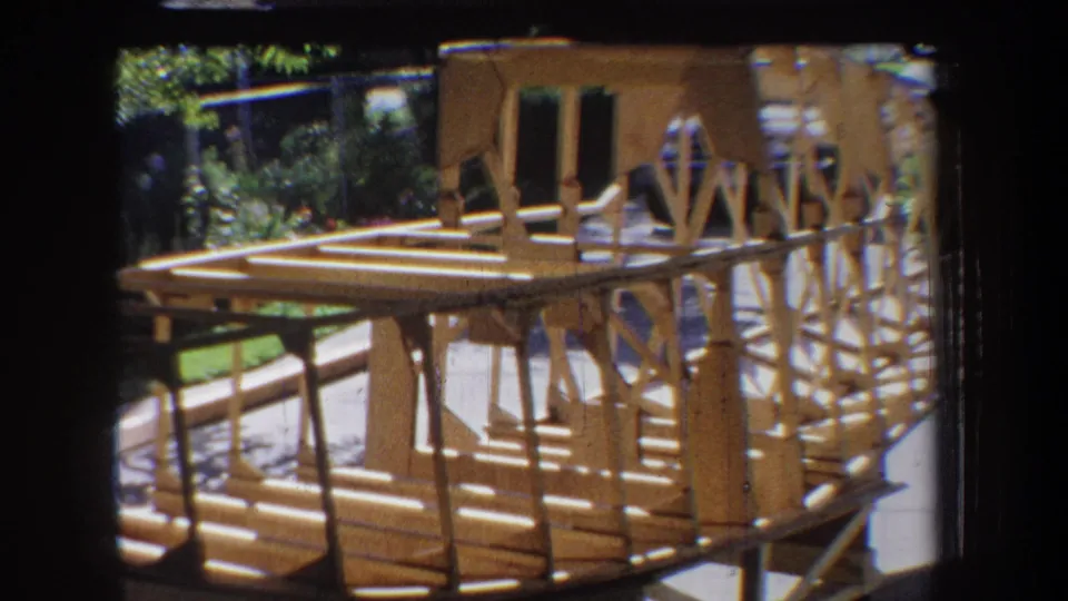 Late-frame 1965 Yucaipa view of the wooden model structure in sunlight, showing its platform, rail-like truss sides, and garden setting.