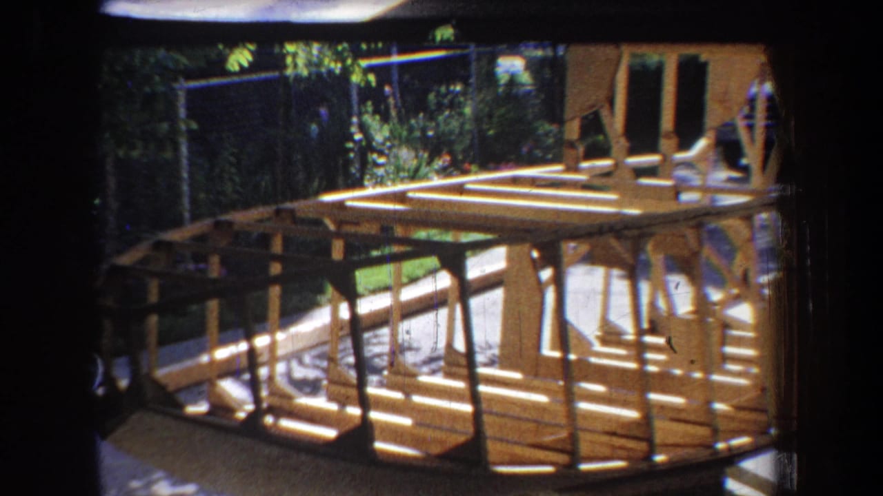 Wooden boat construction continues in the 1965 California home-movie frame, with ribs and planking visible in sharp sun.