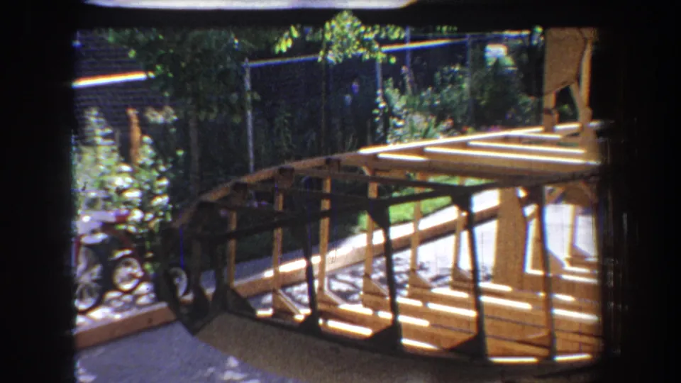 A 1965 Yucaipa home-movie frame shows the backyard boat project from the side with frame lines, supports, and foliage visible.