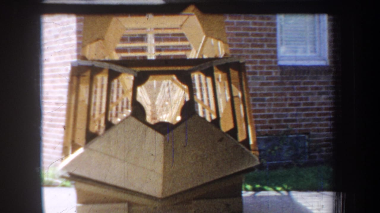 1965 Yucaipa-area 8mm frame showing a faceted wooden sculpture with slatted panels near a brick wall and window.