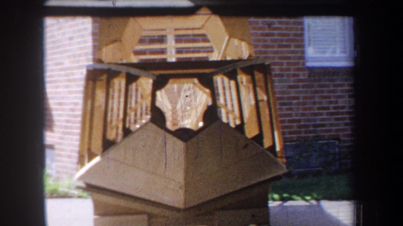 Wooden faceted object with slats viewed in front of brickwork.