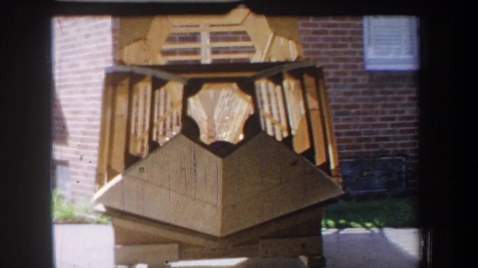 Final 1965 Yucaipa-area frame with the geometric wooden sculpture centered, showing slats, panels, brickwork, and window detail.