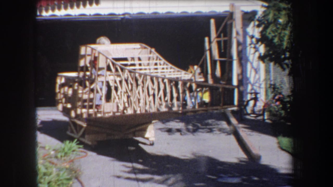 Wooden model structure on a sunny driveway, with a garage opening and yard plants nearby.
