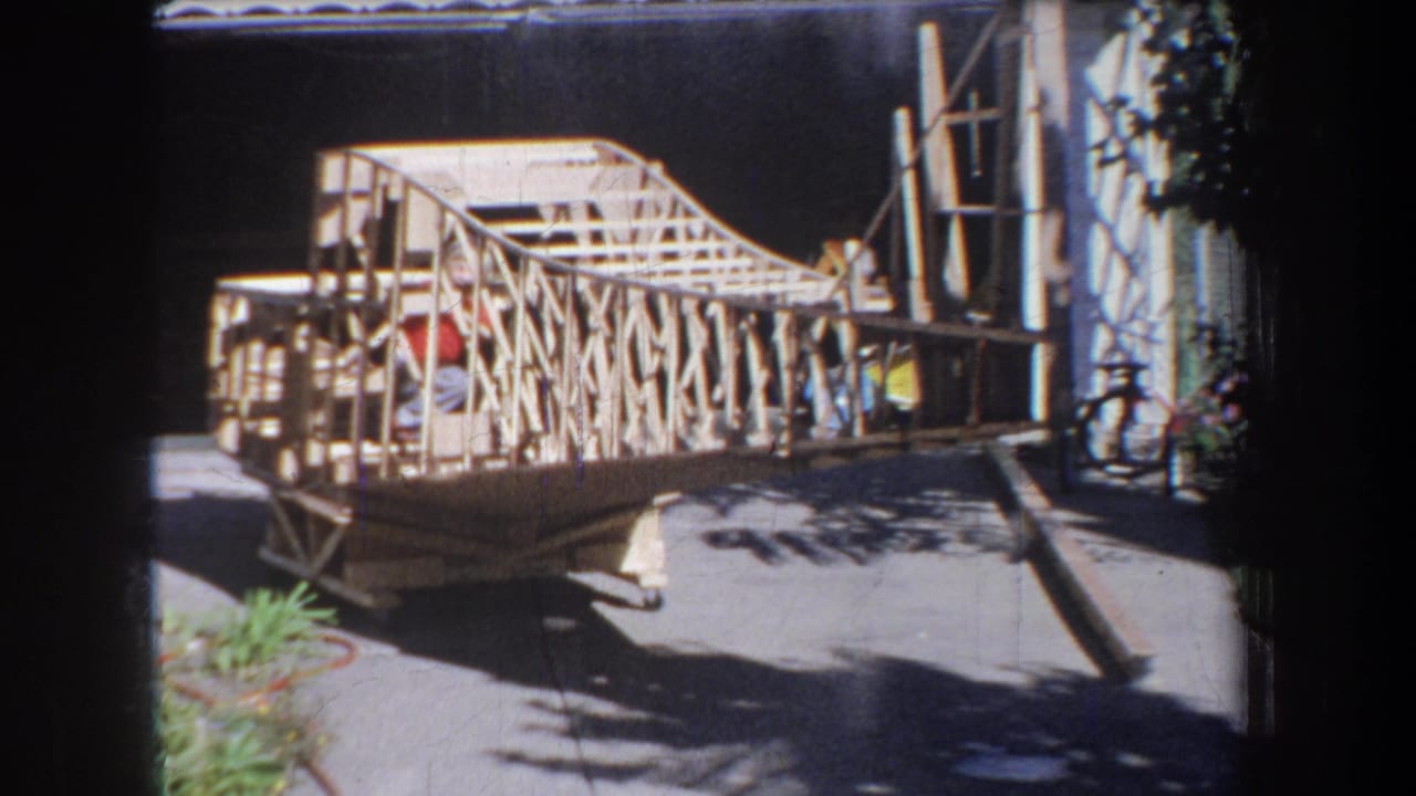 1965 Yucaipa home-movie frame showing a skeletal wooden home-built airplane on a sunny driveway near a house.
