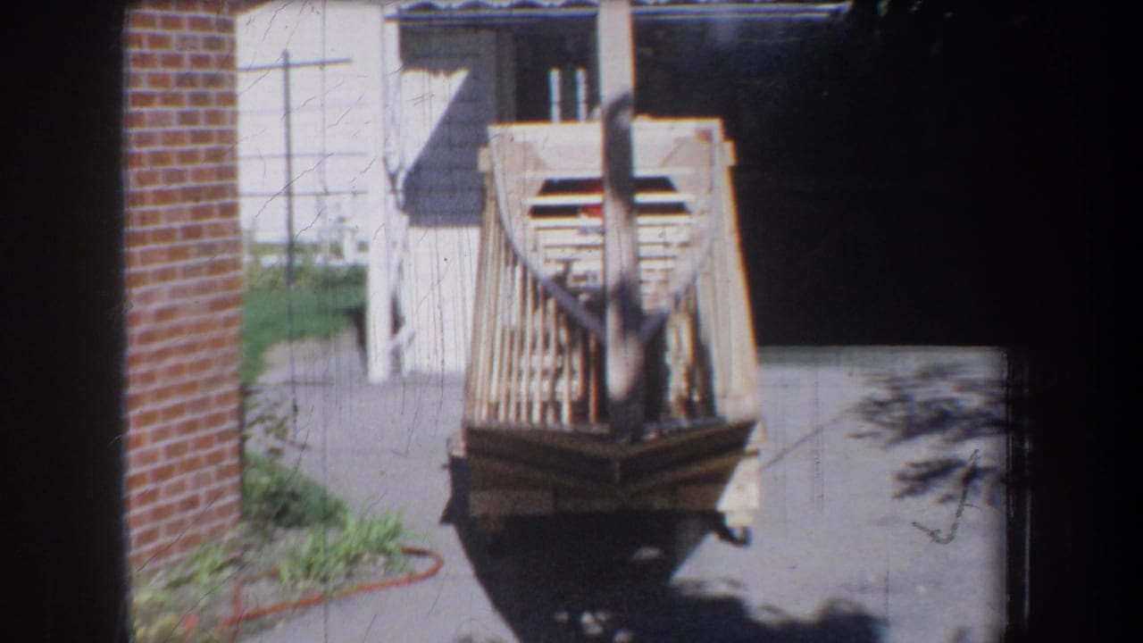 1965 Yucaipa-area home-movie frame showing a light wooden boat frame on a cart beside a garage and brick wall.