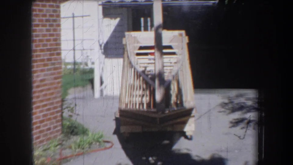 1965 Yucaipa-area home-movie frame showing a light wooden boat frame on a cart beside a garage and brick wall.