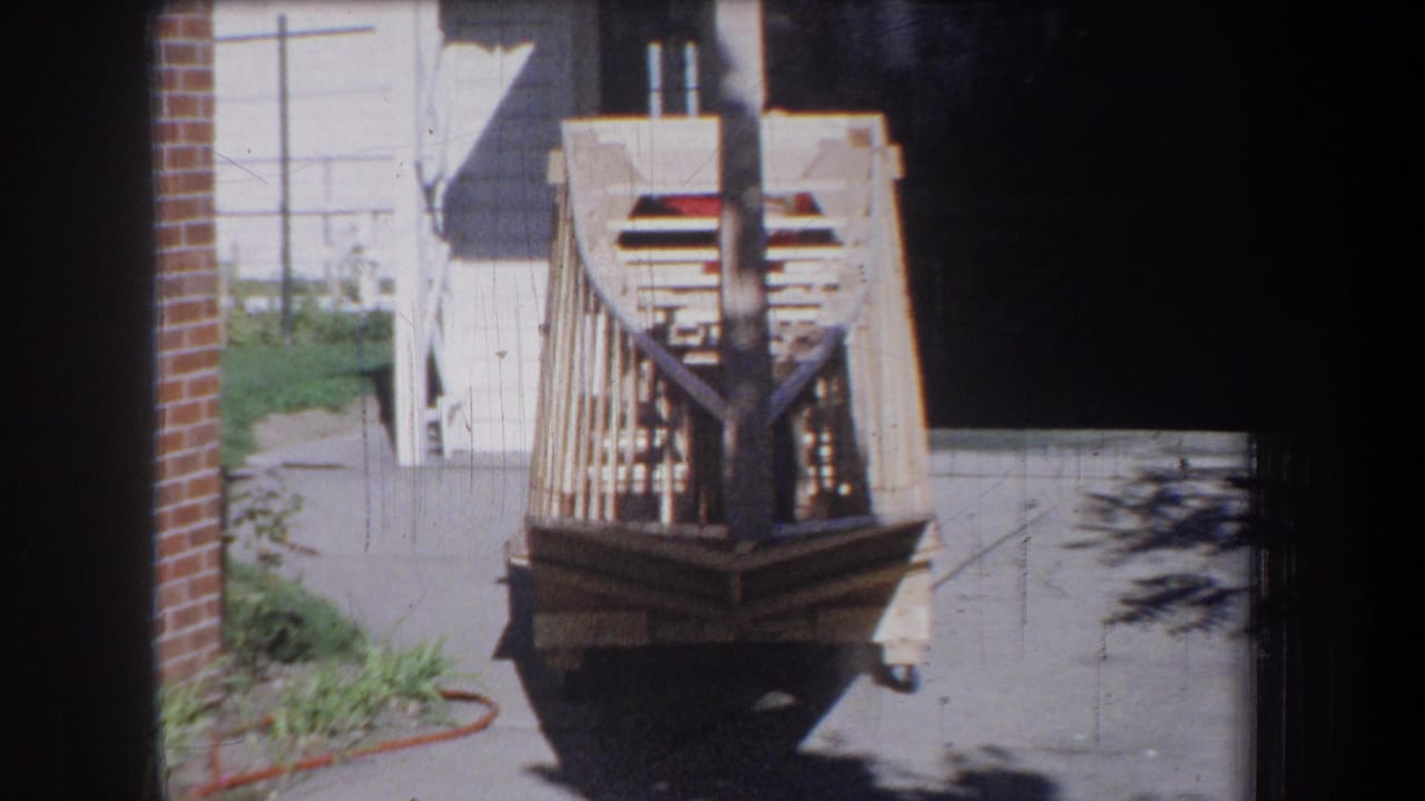 Rear view of a wooden boat frame on a cart outside a garage in daylight.