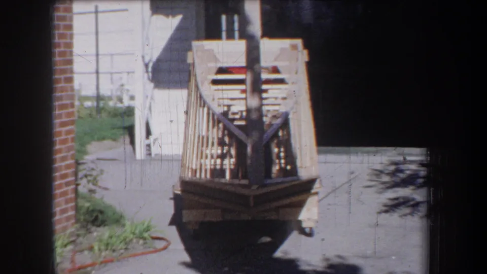 1965 Yucaipa-area frame with the unfinished wooden boat centered on a driveway near white siding and an open garage area.