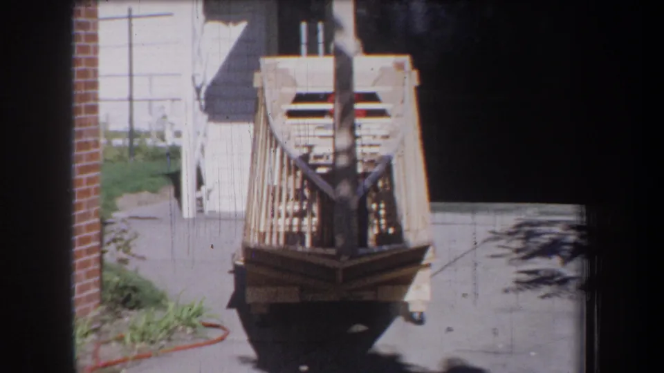1965 Yucaipa-area home-movie frame showing the boat project slightly closer, with exposed woodwork and driveway shadows.