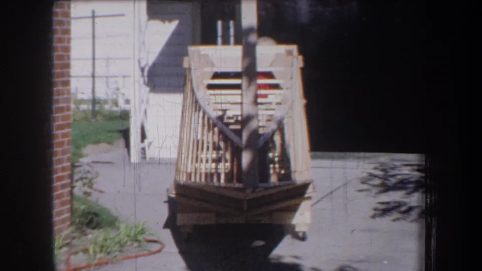 1965 Yucaipa-area frame showing the unfinished boat from the rear, with a dark garage opening and sunlit driveway nearby.