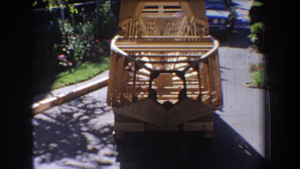 1965 home-movie frame showing the rear of the handmade wooden toy on a driveway with shrubs and a parked car visible.