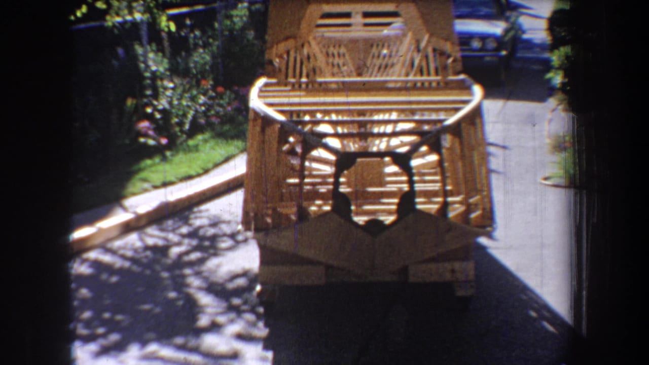 1965 Yucaipa home-movie frame showing a wooden slat structure near pavement, with greenery, shadows, and part of a parked car behind it.