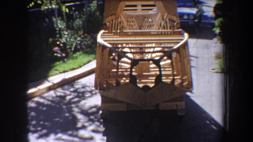 1965 Yucaipa home-movie frame showing a wooden slat structure near pavement, with greenery, shadows, and part of a parked car behind it.