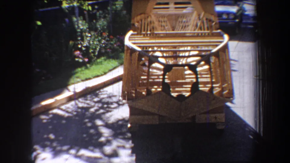 Final 1965 Yucaipa frame with the wooden structure centered near pavement, bordered by garden foliage, shadowed foreground, and a partly visible blue car.