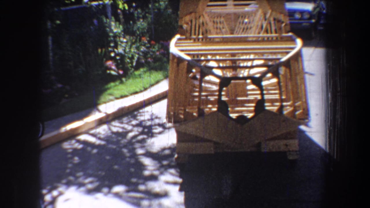 1965 home-movie view of the wooden aircraft-like frame from the front, with driveway, garden plants, and a blue car in the background.