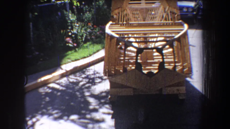 1965 home-movie view of the wooden aircraft-like frame from the front, with driveway, garden plants, and a blue car in the background.
