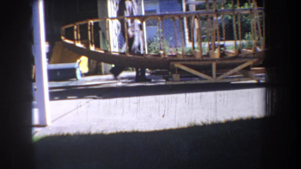 1965 Yucaipa-area home-movie frame showing the side of the wooden fuselage structure near house siding, porch railings, and driveway pavement.