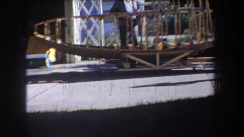 1965 home-movie still showing a side view of the wooden aircraft-style frame, with house details, driveway, and yard shadows visible.