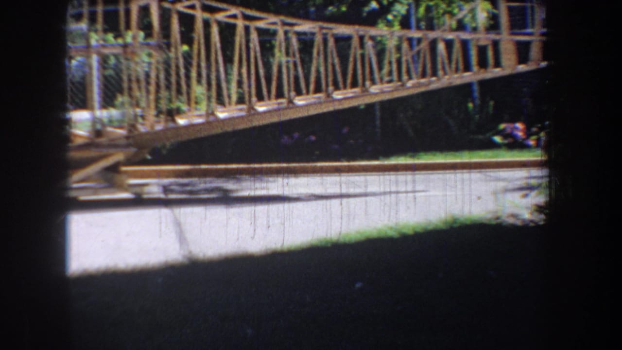 1965 Volmer, Yucaipa home-movie frame showing a metal truss bridge above a paved road, with chain-link fencing and trees behind it.