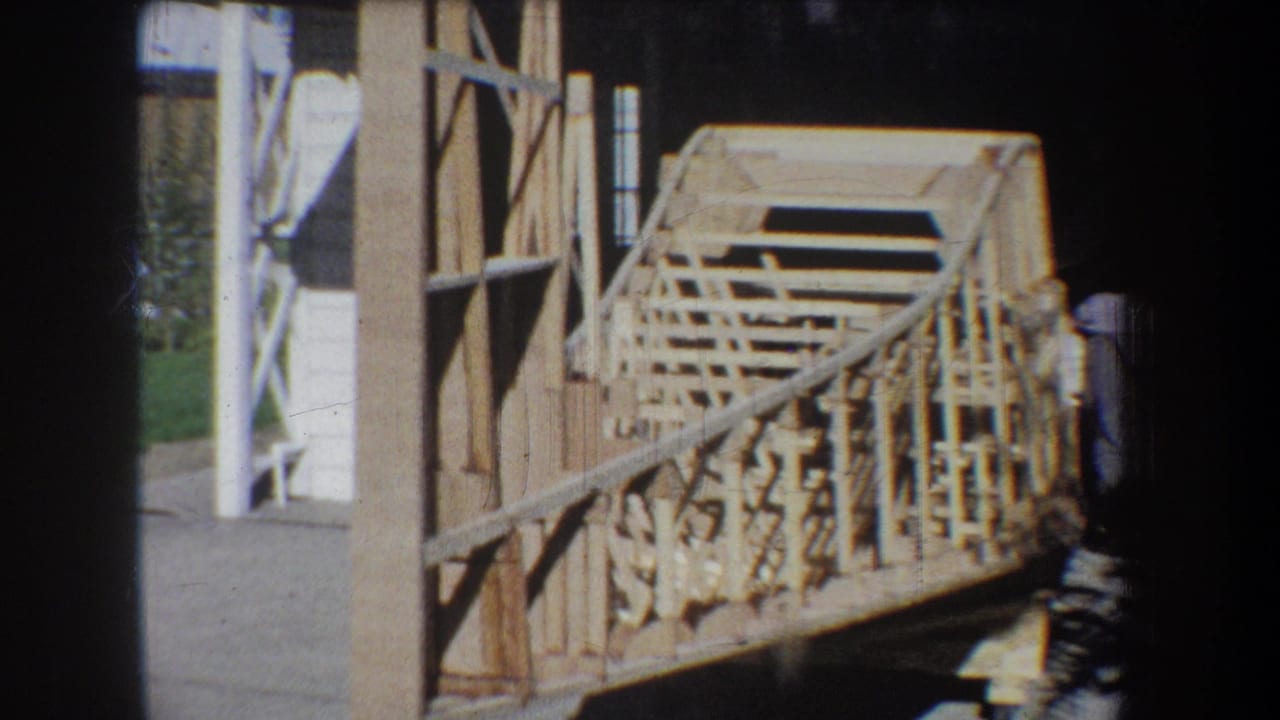 1965 Yucaipa home-movie frame showing a large wooden lattice model near an open garage, with fence, lawn, and a partly visible worker.