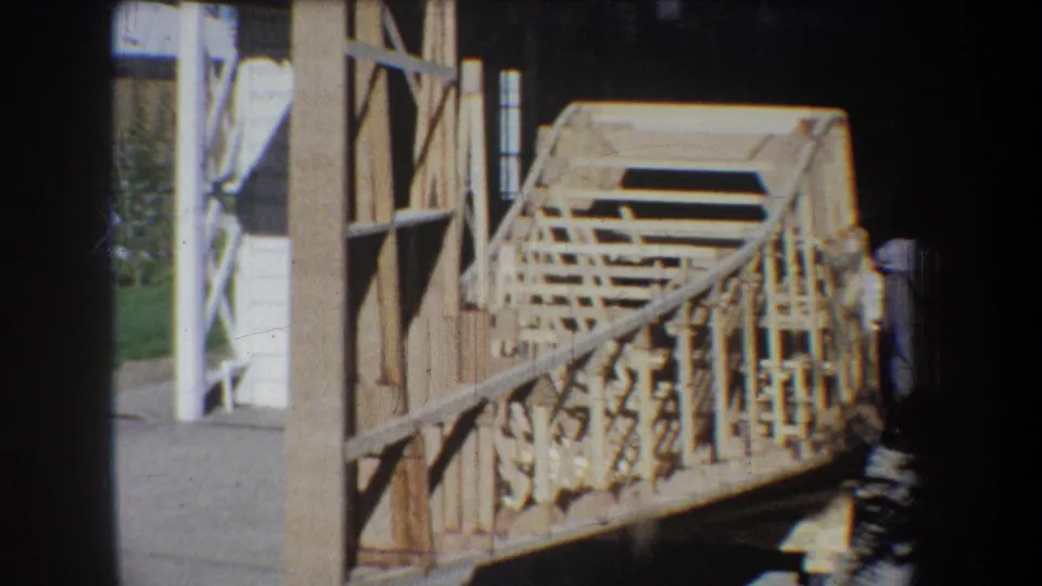 1965 Yucaipa home-movie frame showing a large wooden lattice model near an open garage, with fence, lawn, and a partly visible worker.