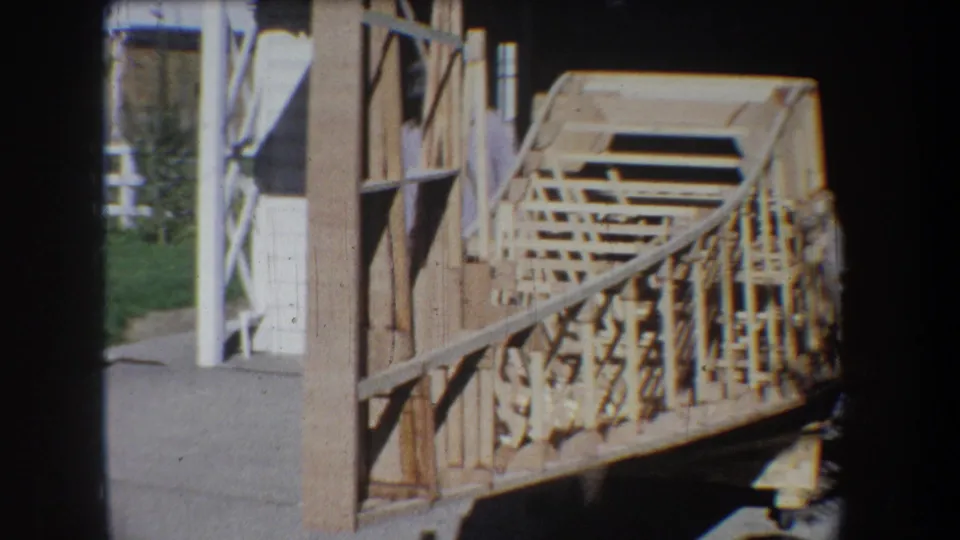 1965 Yucaipa home-movie still showing a side view of a large wooden lattice model extending from a garage area toward the driveway.