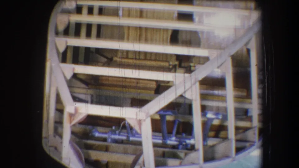 The camera frames the boat structure more closely, emphasizing wooden crosspieces, ribs, and installed blue seats.