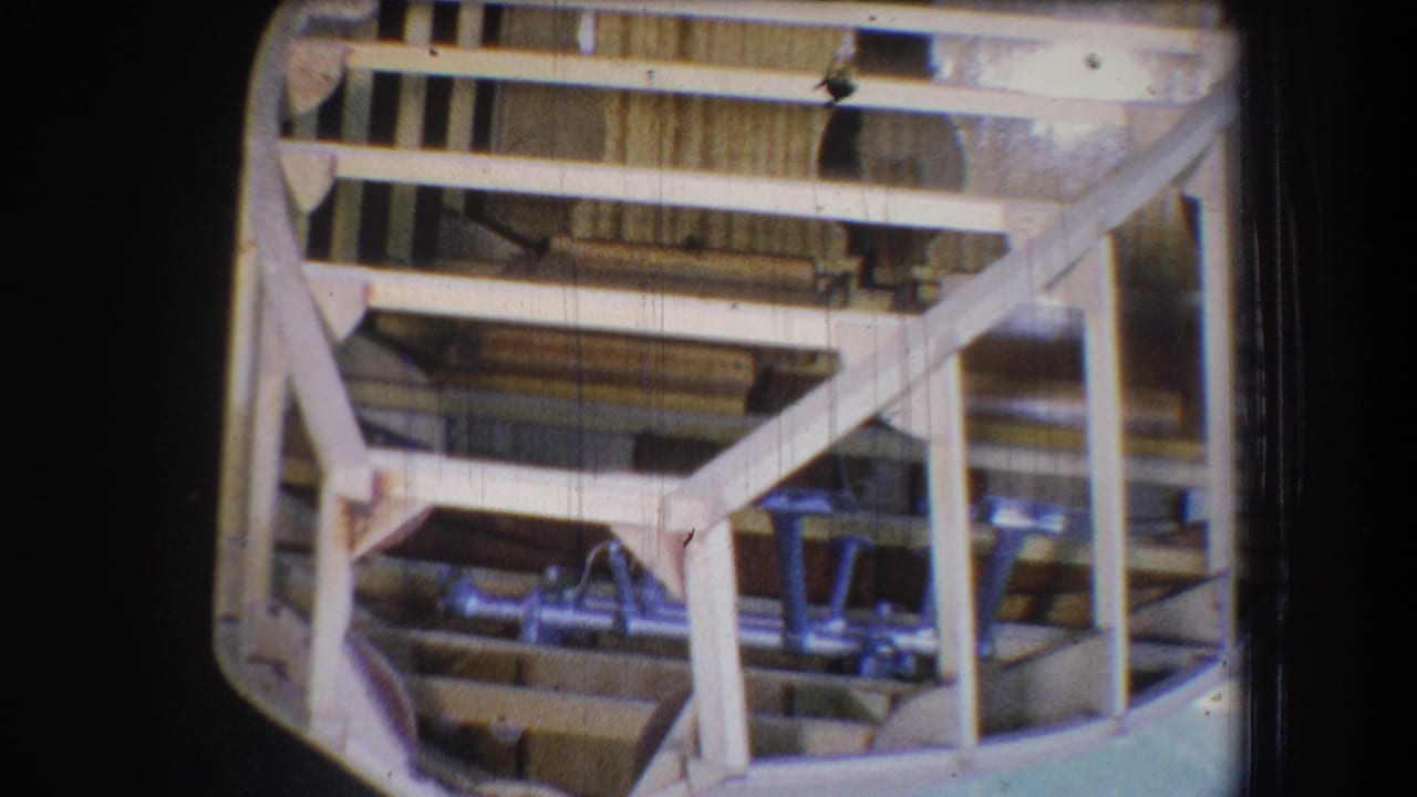 Wooden boat frame with pale beams and metal fixtures inside a dim workshop.
