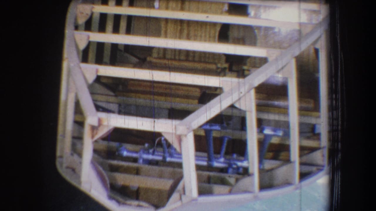 A steady 8mm view shows the unfinished wooden boat frame, angled supports, and metal components in a dim California workshop setting.