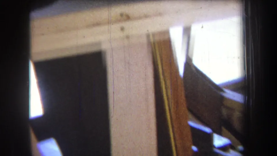 Tilted 8mm frame from the 1965 construction clip showing pale beams, shadowed gaps, and bright light through an opening.
