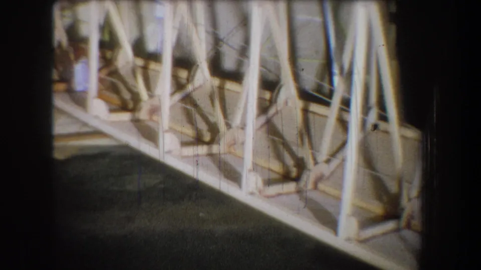 1965 home-movie frame showing the lower portion of a wooden truss, with pale angled members and bracing crossing above a dark floor.
