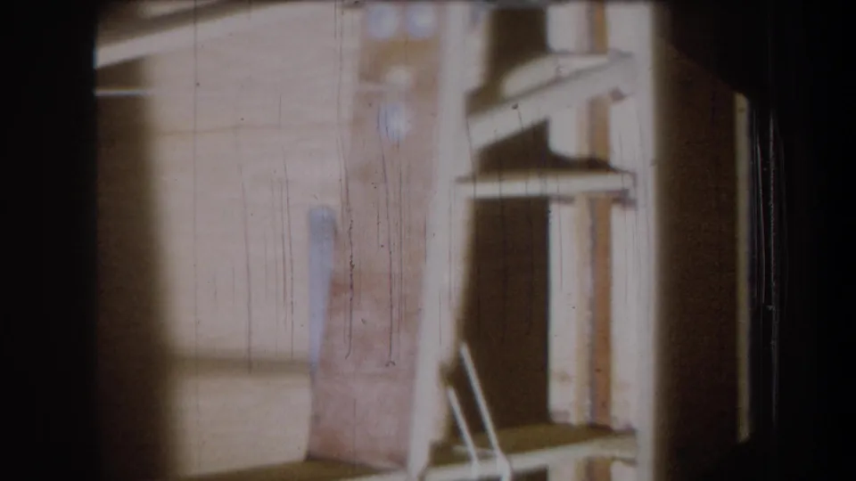 1965 Yucaipa home-movie frame holding on a wooden garage interior, with stored structure details partly obscured by scratches and shadow.