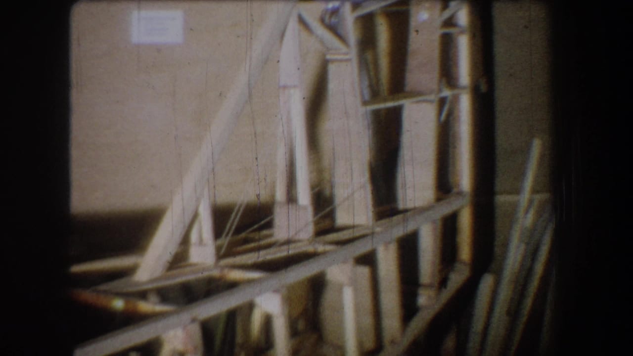 1965 Yucaipa 8mm frame showing a wooden model with angled supports, posts, and a small wall label in a dim indoor setting.