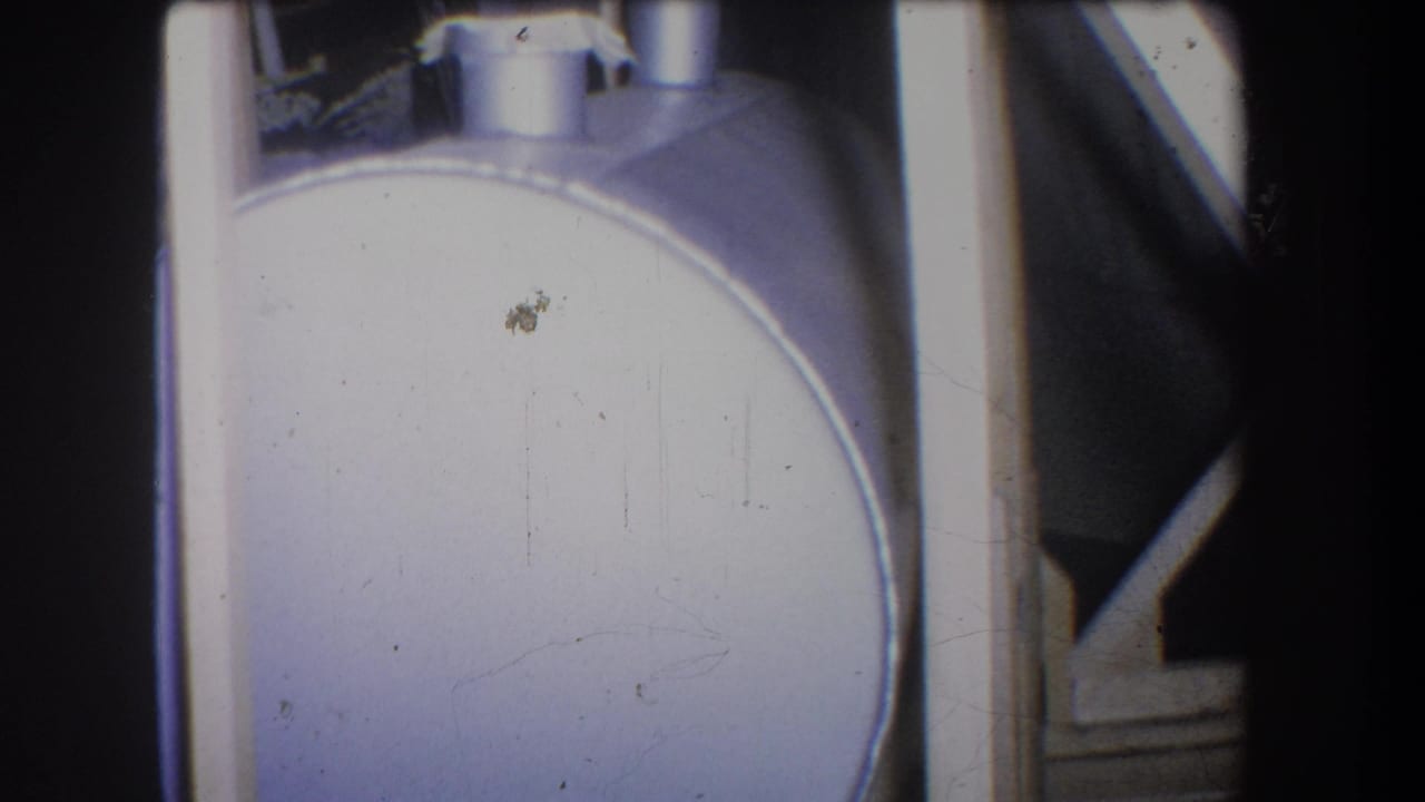 1965 home-movie frame showing a pale cylindrical metal container with vertical support elements and partial fixtures nearby.