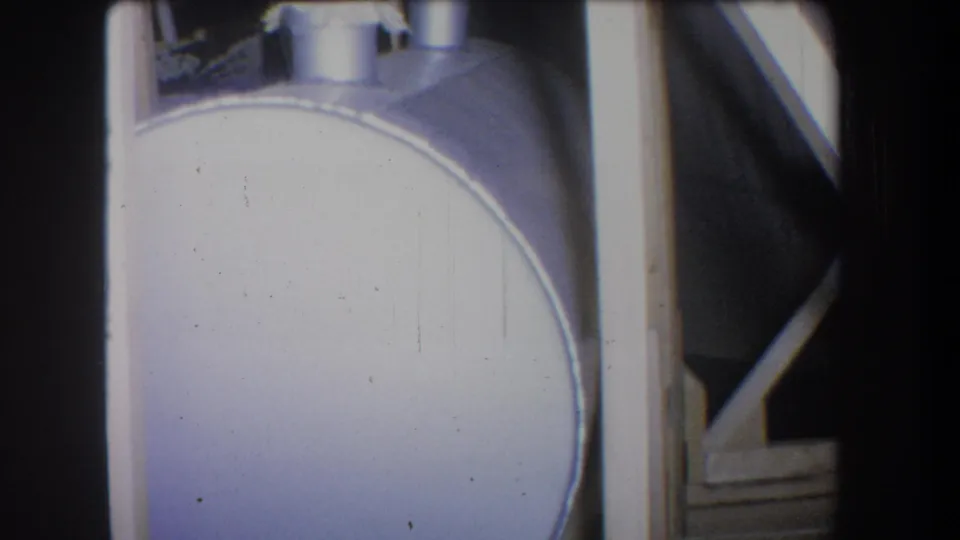 1965 8mm view of the curved side of a large pale metal container with adjacent structural supports and edge shadowing.
