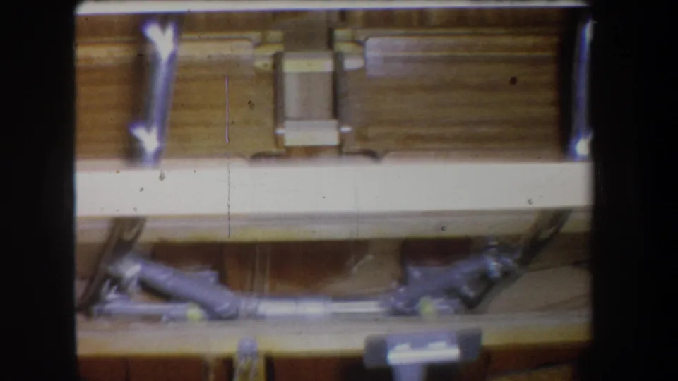 1965 home-movie frame showing wood siding or beams with metal brackets, exposed pipework, and utility fixtures below.