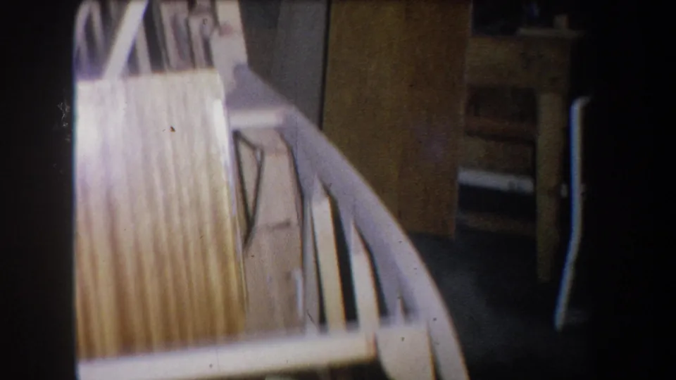 1965 Yucaipa-area home-movie frame showing curved boat ribs, pale planking, and shadowed workshop space around the hull.