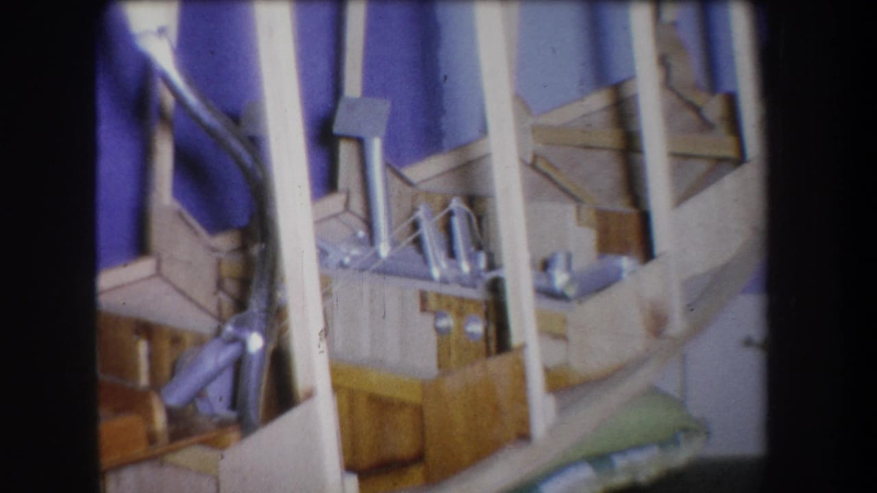 Supported 1965 Yucaipa frame showing a close detail of a wood-and-metal homemade device, with rods and small parts set before purple fabric.