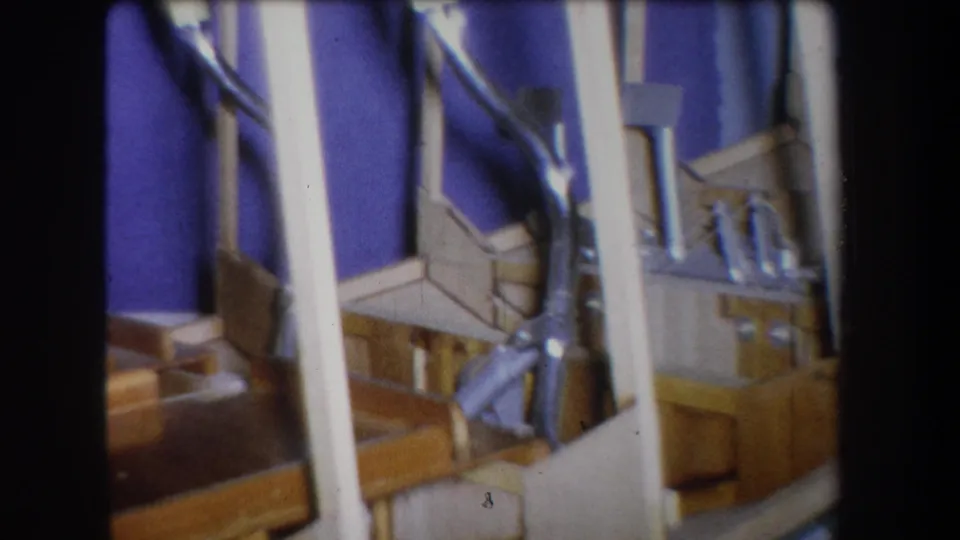 Final 1965 Yucaipa frame showing the homemade wood-and-metal object from a slightly shifted angle, with a curved tube near white supports.