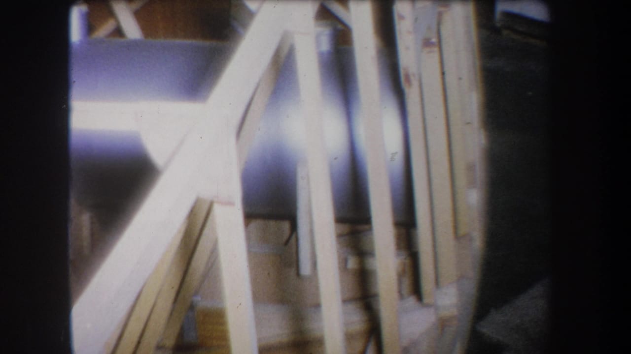 1965 Yucaipa home-movie frame showing pale angled wood supports, vertical framing, and a large bluish metal pipe or tank in soft focus.